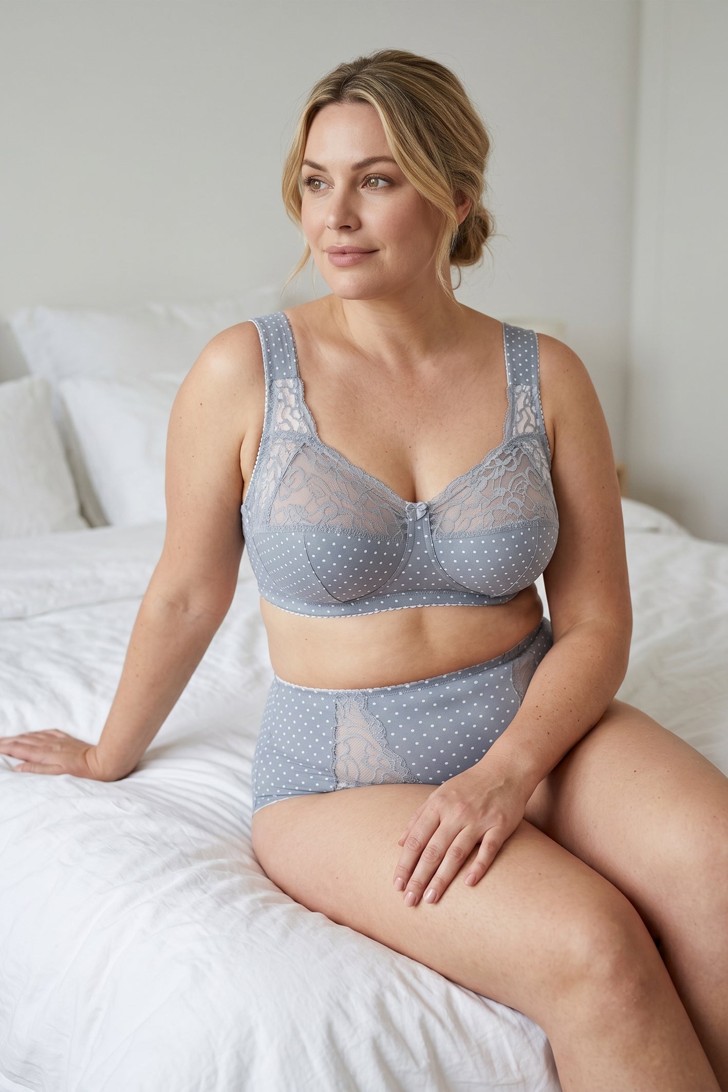 A woman sits on a bed wearing a matching light blue-gray lace and polka dot plus size cotton lingerie set.