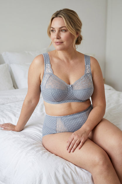 A woman sits on a bed wearing a matching light blue-gray lace and polka dot plus size cotton lingerie set.