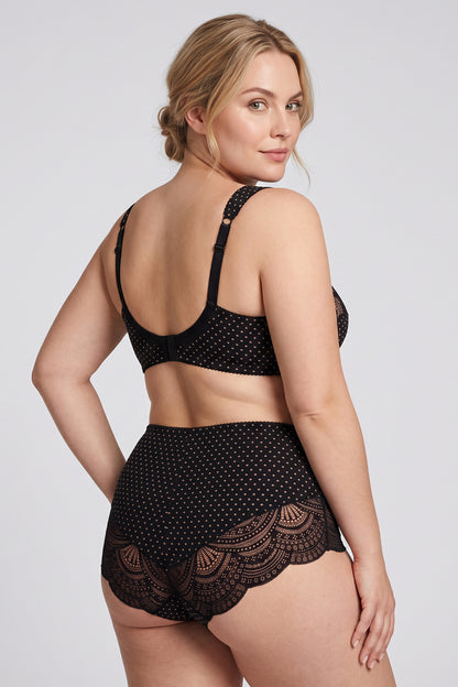A woman modeling a black two-piece cotton lingerie set with lotus polka dots and lace details, shown from a back-three-quarter angle against a light gray studio background.