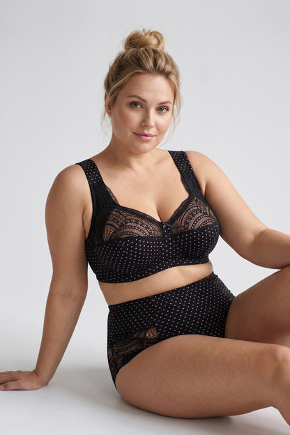A woman wearing a black lace and polka-dot cotton bra and matching high-waisted briefs, seated against a light gray studio background.